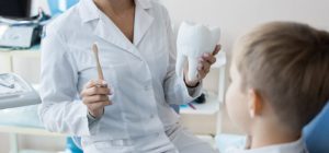 Mid section portrait of female dentist holding toothbrush and tooth model explaining oral hygiene rules to little boy sitting in dental chair